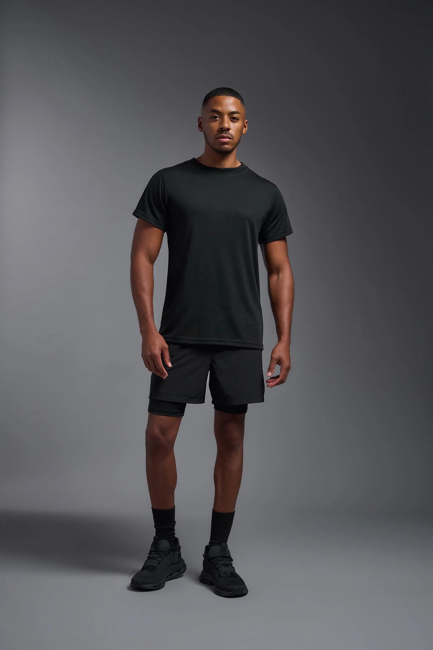 Man wearing a black t-shirt, shorts, and shoes on a gray background