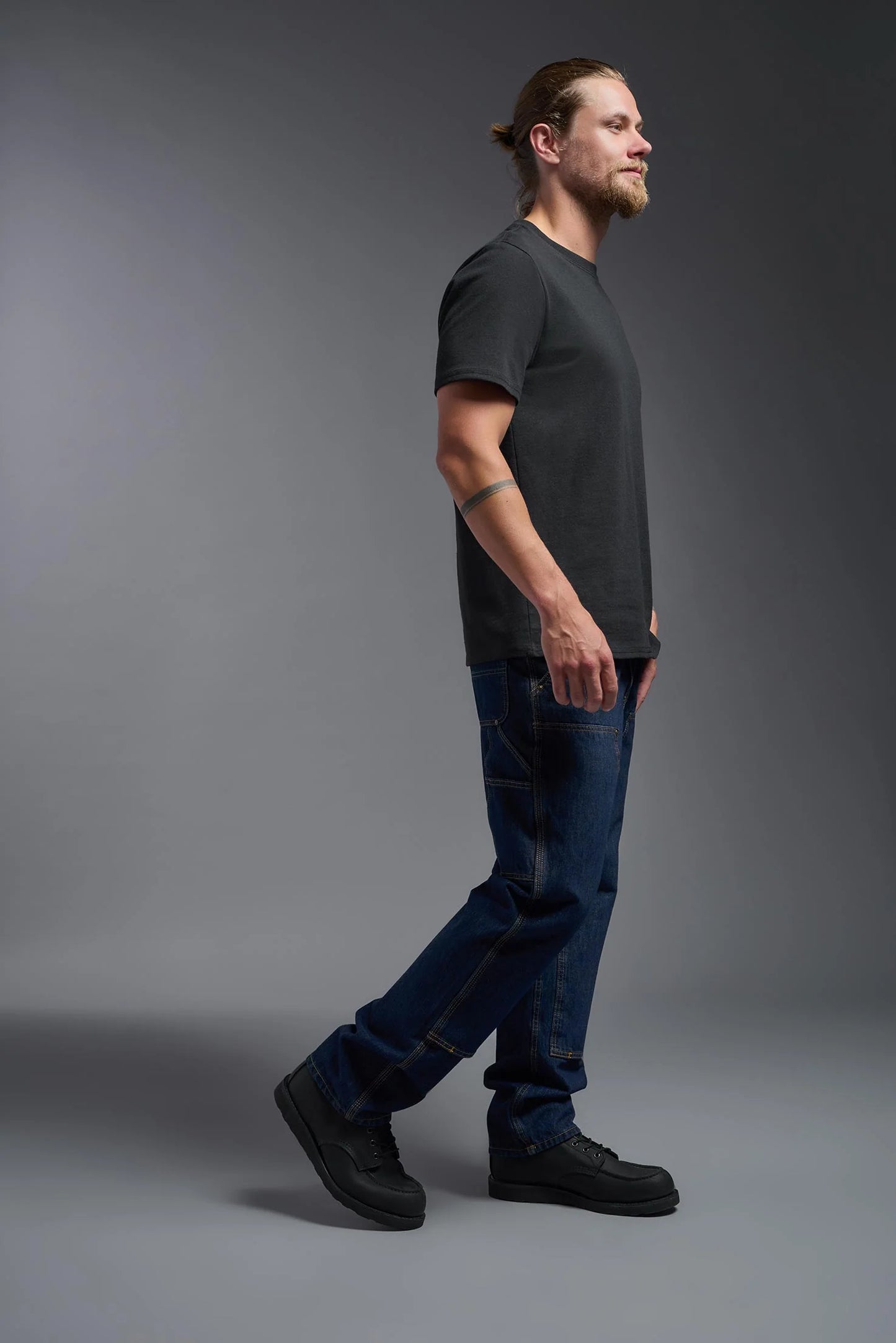 Man wearing a dark gray t-shirt and blue jeans standing against a gray background