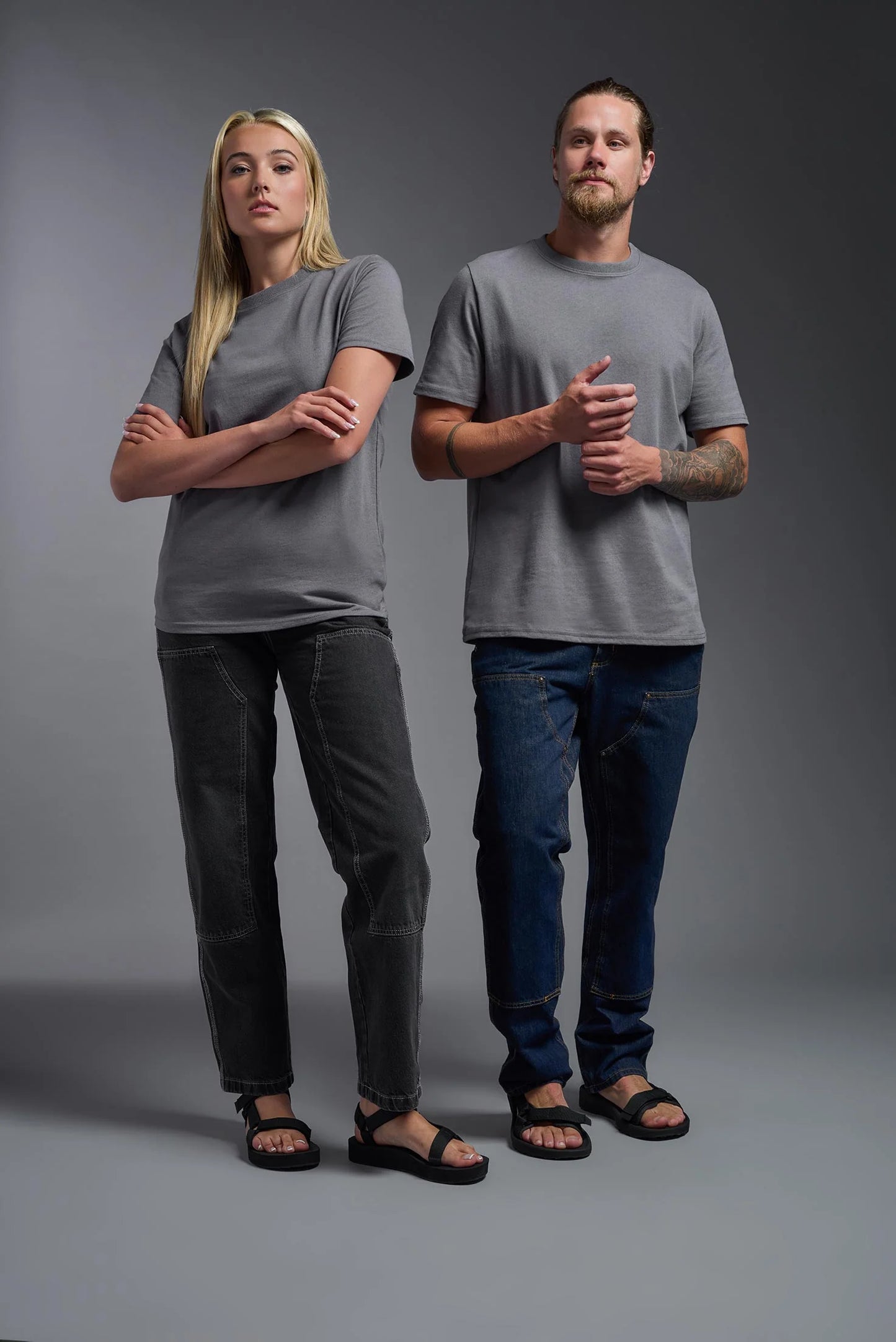 Two people wearing gray t-shirts and dark jeans standing against a gray background