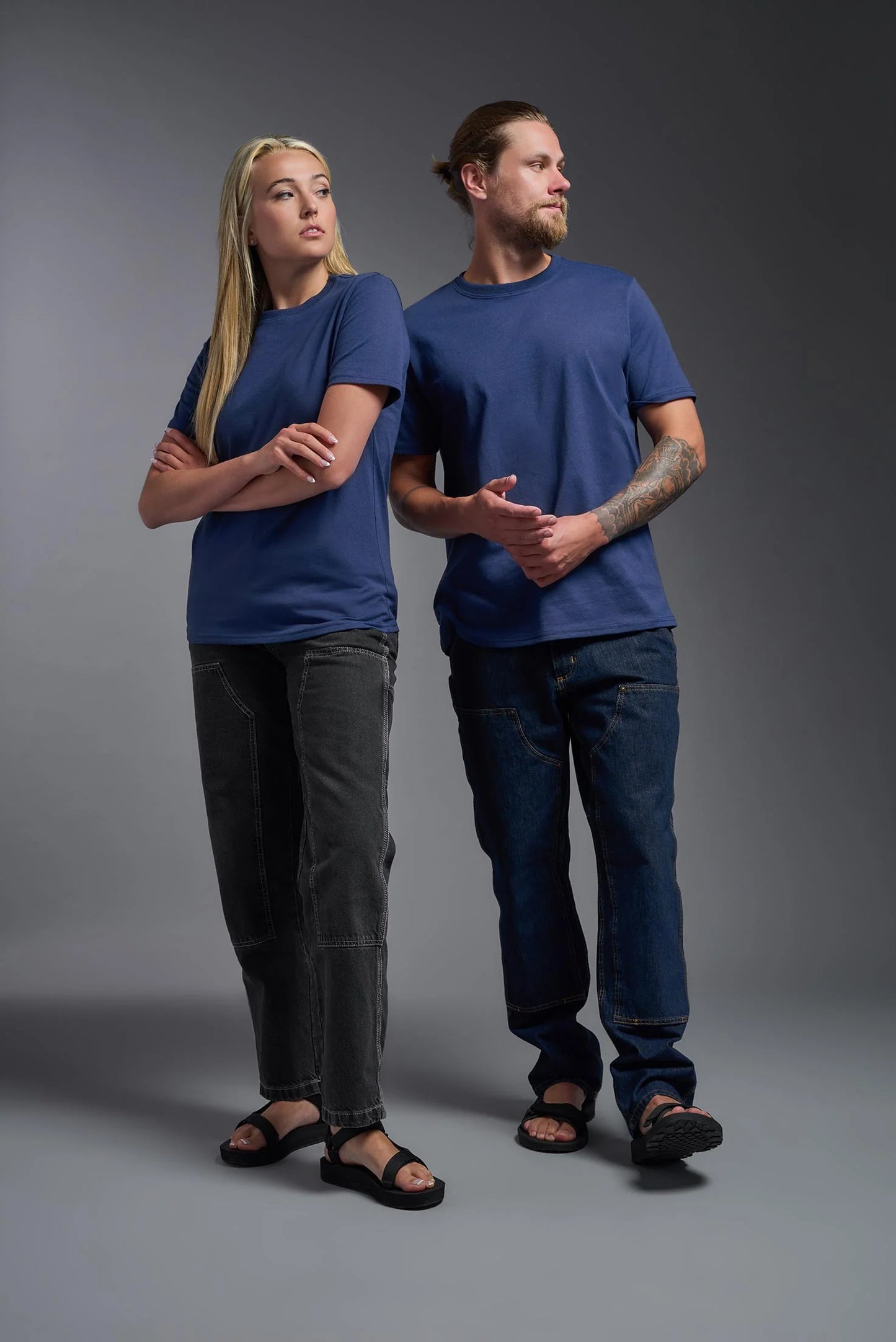 Two people wearing blue t-shirts and dark pants standing against a gray background