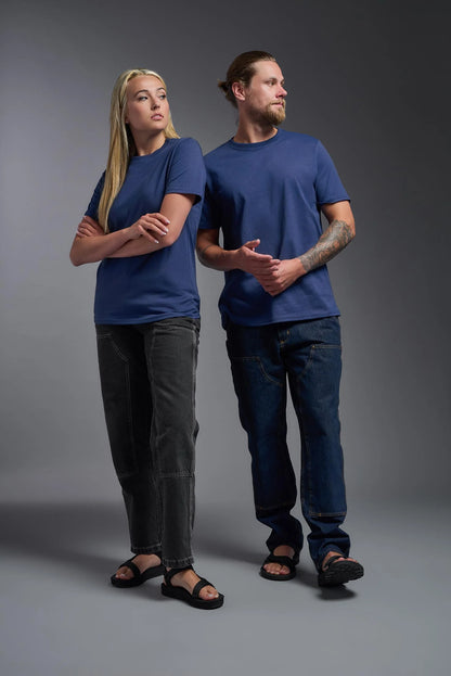 Two people wearing blue t-shirts and dark pants standing against a gray background