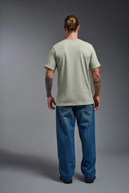 Man wearing a light green t-shirt and blue jeans standing against a gray background