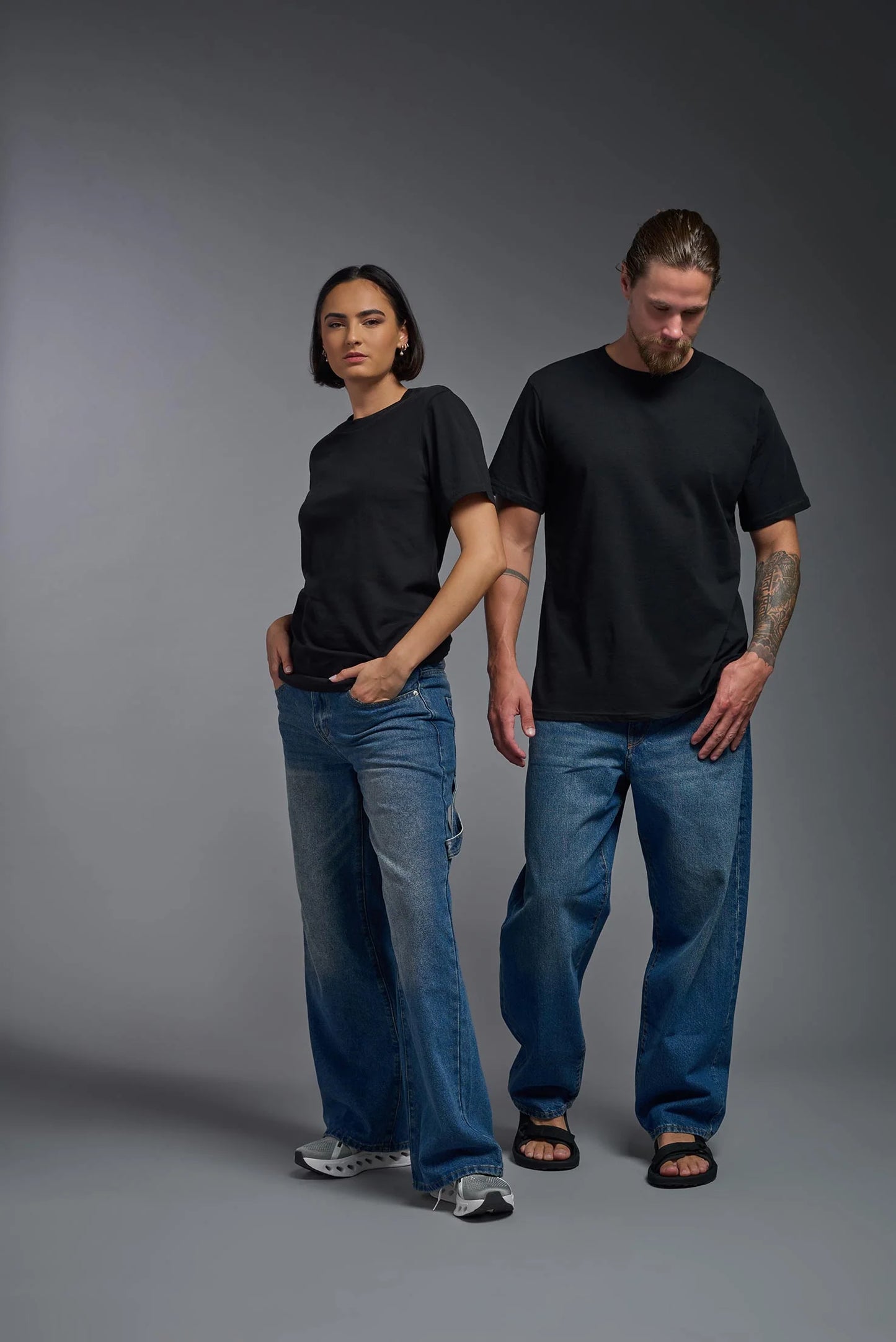 Two people wearing black t-shirts and blue jeans standing against a gray background