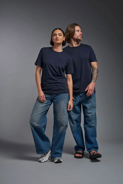 Two people wearing navy t-shirts and blue jeans standing against a gray background