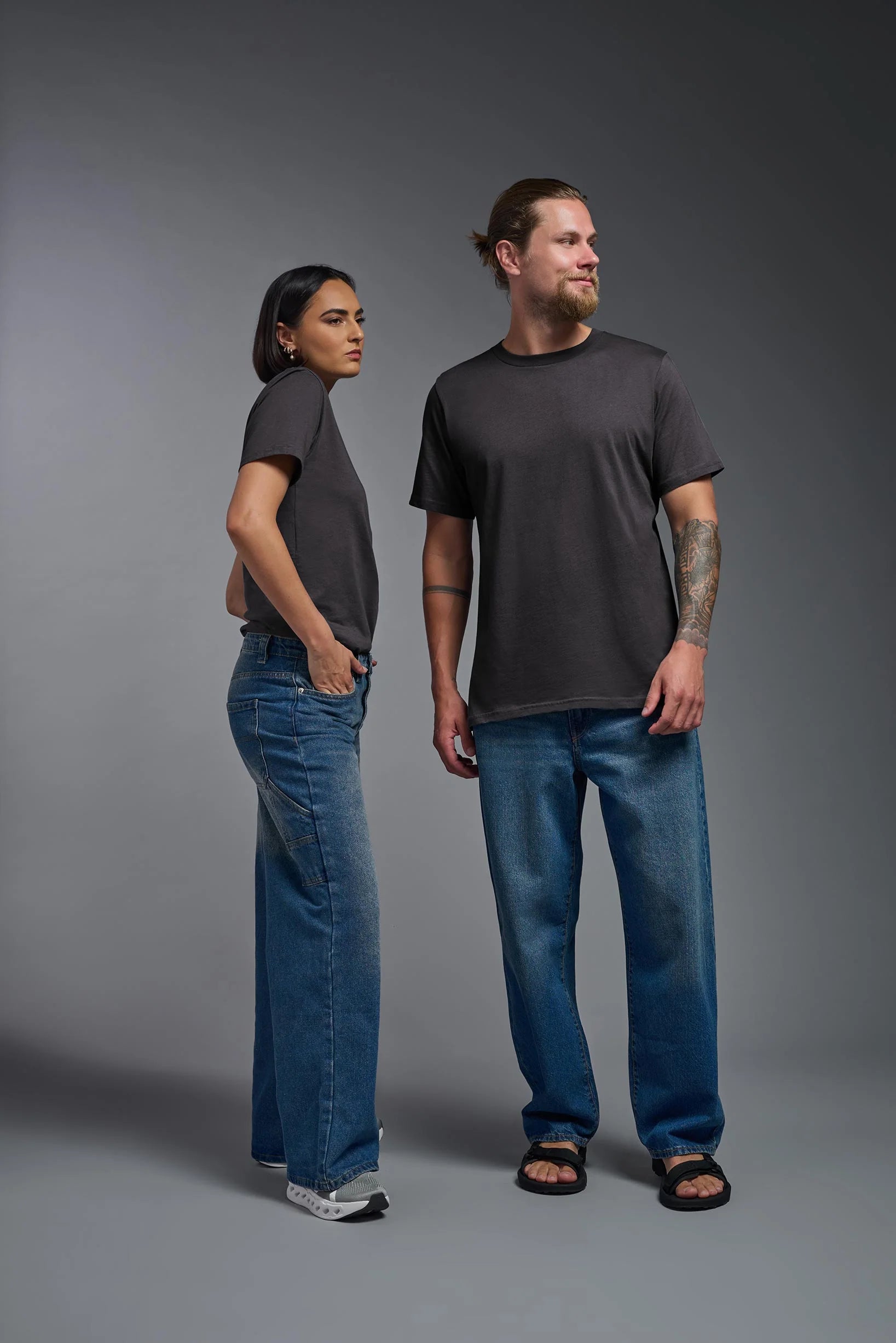 Two people wearing black t-shirts and blue jeans standing against a gray background