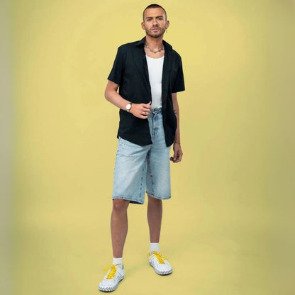 Man wearing a black shirt, white t-shirt, light blue shorts, and yellow sneakers on a yellow background