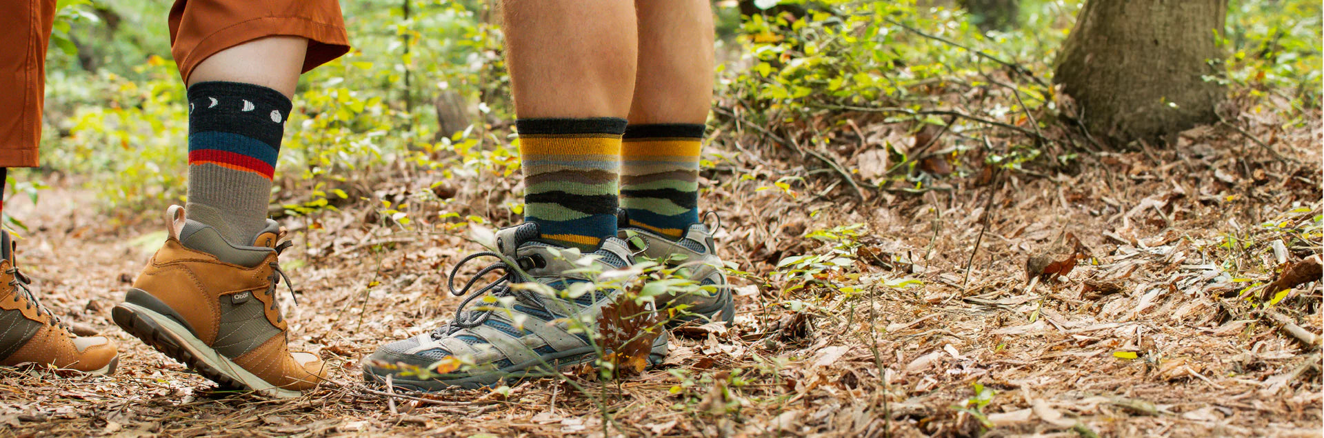 Two pairs of feet wearing hiking boots and colorful socks standing on a forest floor.