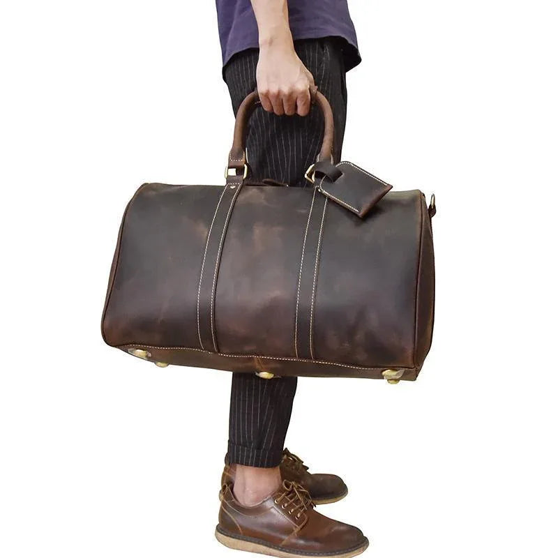 Brown leather duffel bag with top handles, shoulder strap, and metal corner feet