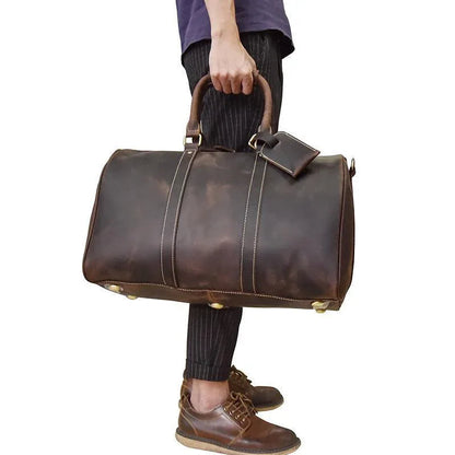 Brown leather duffel bag with top handles, shoulder strap, and metal corner feet